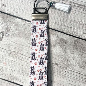 May include: White and red patterned key chain with a black and white dog design. The key chain has a silver metal ring and a white tassel.