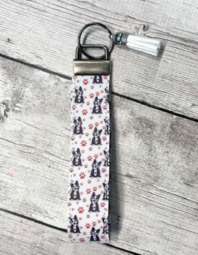 May include: White and red patterned key chain with a black and white dog design. The key chain has a silver metal ring and a white tassel.