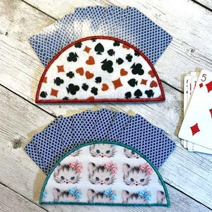 May include: Two half-circle playing card holders with card suit designs and kitten patterns. The top holder has red, black, and white card suits with a red border. The bottom holder has a kitten pattern with pink and blue bows and a teal border.