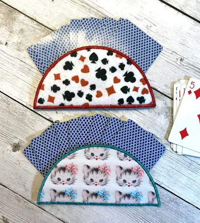 May include: Two half-circle playing card holders with card suit designs and kitten patterns. The top holder has red, black, and white card suits with a red border. The bottom holder has a kitten pattern with pink and blue bows and a teal border.
