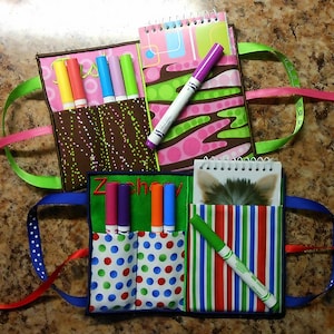 Könnte beinhalten: Handgefertigte Stoff-Bastelutensilien-Organizer mit bunten Markern und Notizbüchern. Ein Organizer hat einen braunen Stoff mit weißen Punkten, ein anderer ein grünes und rosa Design und der dritte einen grünen Hintergrund mit dem Namen "Zachary". Jeder hat Schleifenhenkel.