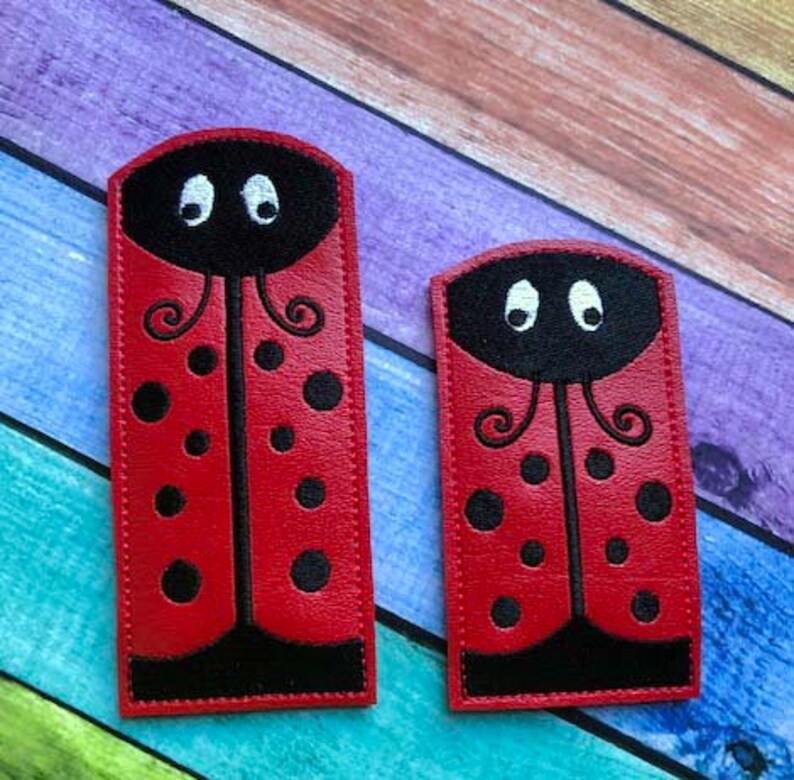 May include: Two red ladybug-shaped patches with black spots and black detailing. The patches have a black head with white eyes and a curved antenna design. One patch is larger than the other.