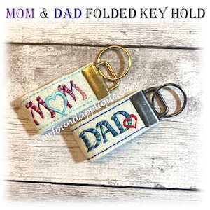 May include: Two folded key holders with the words "MOM" and "DAD" embroidered in colorful thread. The "MOM" key holder features a heart-shaped design. Each key holder has a metal ring for attaching keys. The text "MOM & DAD FOLDED KEY HOLD" is at the top.