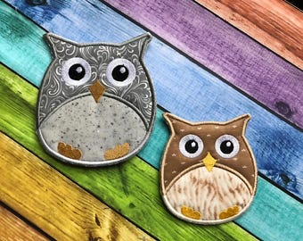 May include: Two felt owl ornaments with embroidered details. The larger owl is gray with a silver swirl pattern and black eyes. The smaller owl is brown with a tan swirl pattern and black eyes. Both owls have yellow beaks.