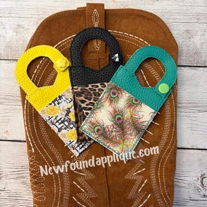 May include: Three decorative keychains with different patterns: yellow with floral print, black with leopard print, and teal with peacock feather print. Each keychain has a handle and is displayed on a brown cowboy boot.
