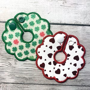 May include: Two decorative, flower-shaped bibs. One is white with green shamrocks and pink accents, a green border, and a green snap. The other is white with brown hearts, a red border, and a white snap.