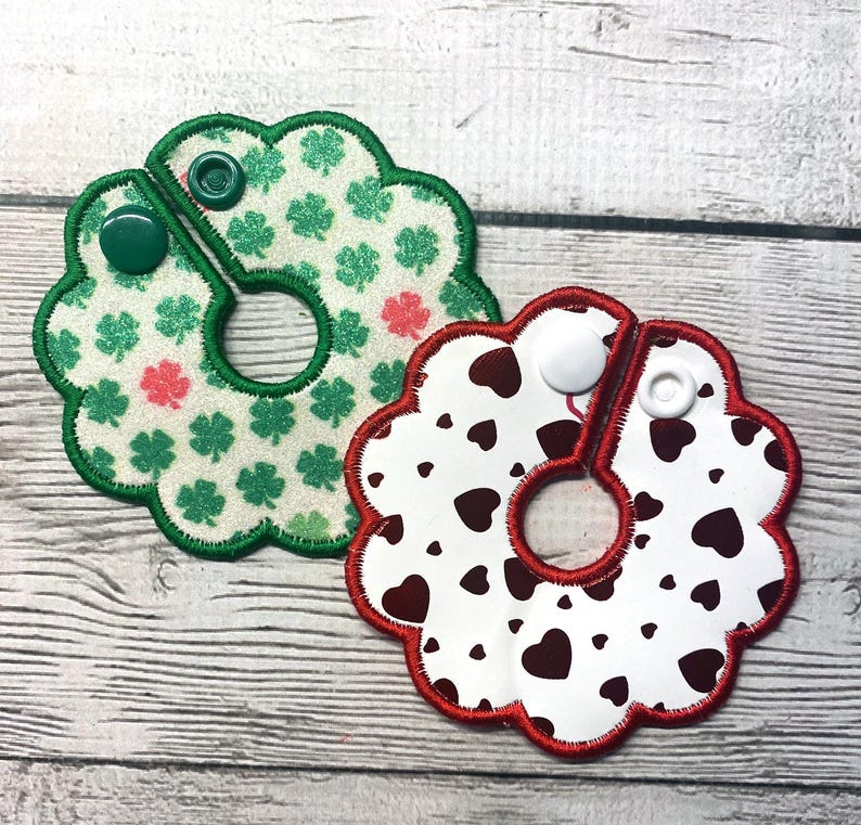 May include: Two decorative, flower-shaped bibs. One is white with green shamrocks and pink accents, a green border, and a green snap. The other is white with brown hearts, a red border, and a white snap.