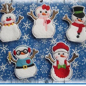 May include: A collection of five felt snowman ornaments with various accessories. Each snowman has a white body, black eyes, and a carrot nose. Accessories include a top hat, earmuffs, a bow, and a scarf. The background is blue with white snowflakes.