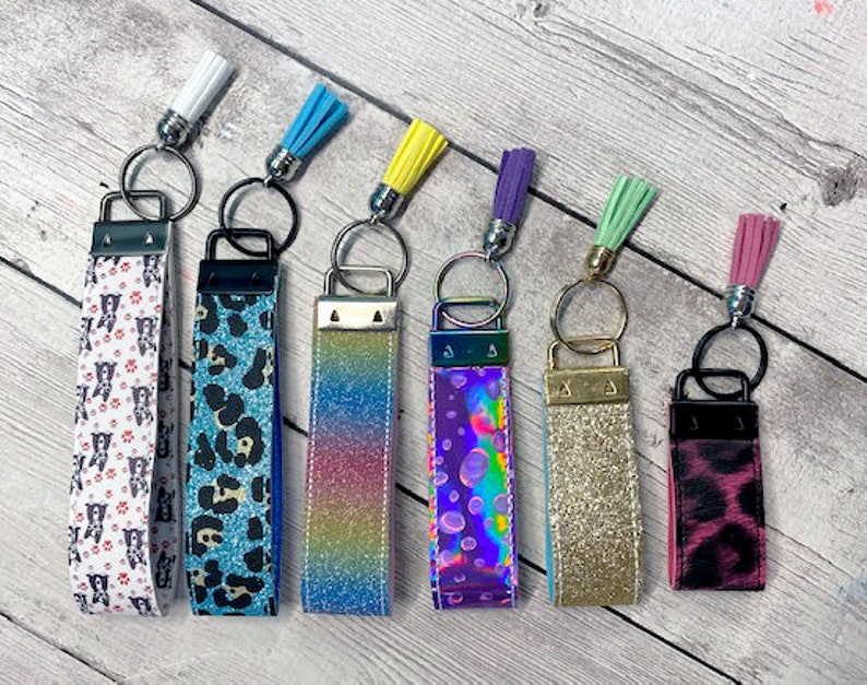 May include: Seven colorful keychains with different patterns and tassels. The keychains are made of fabric and have a metal ring at the top. The patterns include leopard print, glitter, rainbow, and polka dots.