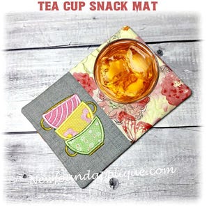 May include: A quilted tea cup snack mat with a glass of iced tea. The mat features a gray section with embroidered stacked tea cups in pink, yellow, and green. The other section has a floral pattern. Text at the top reads "TEA CUP SNACK MAT".