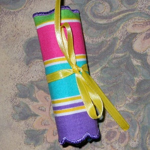 May include: A rolled fabric item with pink, turquoise, yellow, and purple horizontal stripes, tied with a yellow ribbon. The edges are scalloped and trimmed with green. The background has a floral pattern.