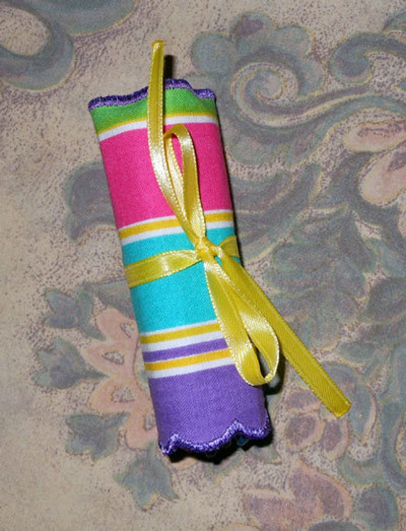May include: A rolled fabric item with pink, turquoise, yellow, and purple horizontal stripes, tied with a yellow ribbon. The edges are scalloped and trimmed with green. The background has a floral pattern.