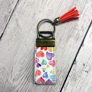 May include: A white keychain with a colorful heart pattern and a gold-colored key ring. A red tassel hangs from the key ring.