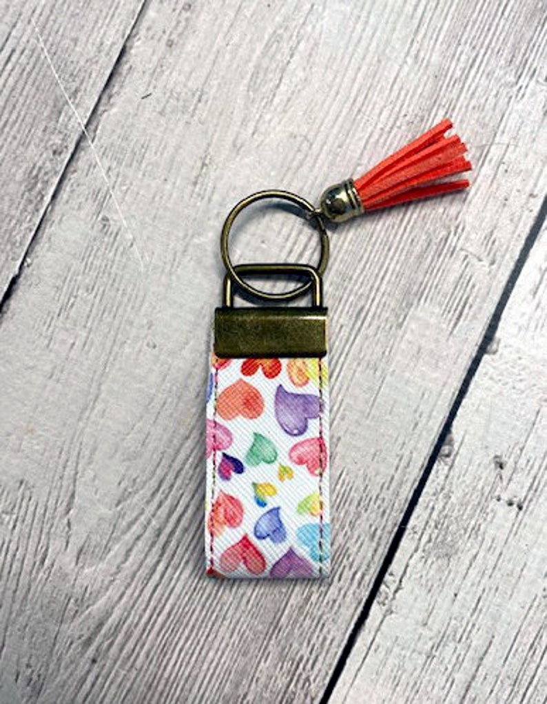 May include: A white keychain with a colorful heart pattern and a gold-colored key ring. A red tassel hangs from the key ring.