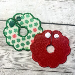 May include: Two decorative, flower-shaped items. One is red, made of textured material, and the other is white with green and pink shamrocks. Both have a snap closure and a circular opening in the center.