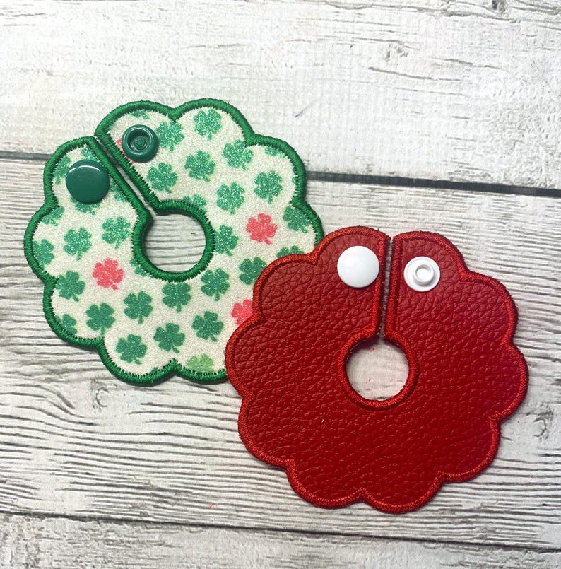 May include: Two decorative, flower-shaped items. One is red, made of textured material, and the other is white with green and pink shamrocks. Both have a snap closure and a circular opening in the center.