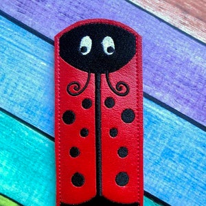 May include: A red ladybug-shaped accessory with black spots and a black head featuring white eyes. The ladybug design is set against a colorful, multi-toned background. The accessory is rectangular with rounded top edges.