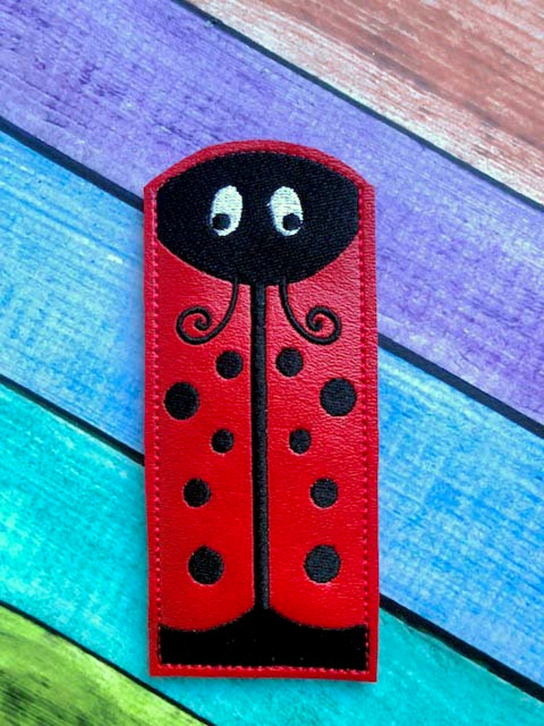 May include: A red ladybug-shaped accessory with black spots and a black head featuring white eyes. The ladybug design is set against a colorful, multi-toned background. The accessory is rectangular with rounded top edges.