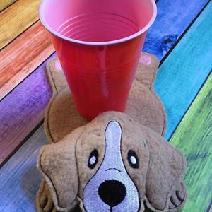 Puede incluir: Un posavasos de fieltro color canela con forma de perro y rasgos faciales bordados, incluyendo un hocico blanco, nariz negra y lengua rosa, sosteniendo un vaso de plástico rosa translúcido. El posavasos tiene orejas caídas y patas.