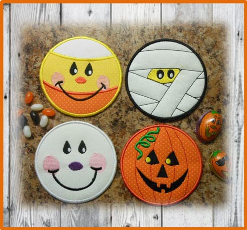 May include: Four round Halloween-themed coasters with embroidered designs. The coasters feature a smiling candy corn, a mummy, a ghost, and a jack-o'-lantern.