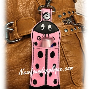 May include: Pink ladybug lip balm holder with black spots and accents, attached to a silver clip. The holder is 5" x 7" and has a pocket holding a lip balm. The text "Lady Bug Lip Balm Holder" and "Newfoundapplique.com" are visible.