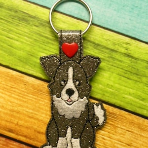Könnte beinhalten: Ein glitzernder schwarz-weißer Hunde-Schlüsselanhänger mit rotem Herzakzent. Der Hund ist gestickt und ein silberner Schlüsselring ist befestigt. Ein niedliches Accessoire für Hundeliebhaber.