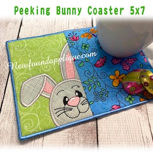 Może przedstawiać: Zielony i niebieski haftowany podkładka pod kubek z królikiem wyglądającym zza zielonego liścia. Podkładka ma kwiatowy wzór z motylami i tekstem "Newfoundlandapplique.com".