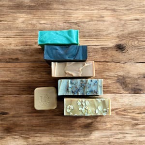 May include: A collection of five rectangular bars of soap and one small square soap bar. The bars include a bright teal, a dark charcoal, a smooth tan, a light beige with dried lavender buds, and a tan bar topped with visible rolled oats.