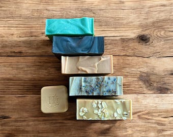May include: A collection of five rectangular bars of soap and one small square soap bar. The bars include a bright teal, a dark charcoal, a smooth tan, a light beige with dried lavender buds, and a tan bar topped with visible rolled oats.