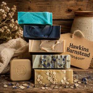 May include: A stack of five rectangular bars of soap in teal, charcoal, tan, and beige, with one bar featuring dried blue petals and another topped with rolled oats. A small square tan soap bar with an HB logo and a tag reading Hawkins Barnstead.