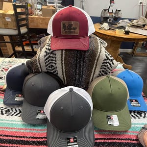 May include: A collection of trucker hats in various colors, including navy, gray, olive green, and blue. One hat features a burgundy front panel with a white mesh back and a leather patch. The hats are displayed on a colorful striped blanket.