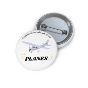 Puede incluir: Botón blanco con alfiler plateado. Presenta un avión pequeño, la palabra "PLANES" en negro y el texto "introverted but will talk about" en una fuente curva. Estrellas doradas adornan el diseño.