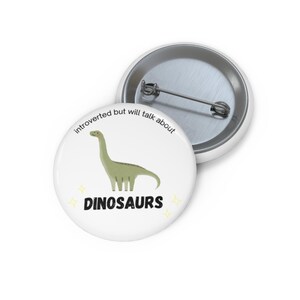Puede incluir: Pin blanco con una ilustración de dinosaurio verde y la palabra "DINOSAURIOS" en negro. El texto "introvertido pero hablará sobre" está impreso alrededor del dinosaurio. El pin es redondo y tiene un respaldo de metal.