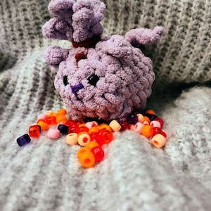 Pode incluir: Uma criatura de pelúcia lavanda feita à mão, com olhos pretos e uma faixa vermelha ao redor do pescoço. A criatura tem um corpo macio e texturizado e um grupo de tufos roxos na cabeça. Contas coloridas em tons de vermelho, laranja e roxo estão espalhadas ao redor.