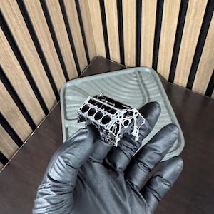 May include: A detailed, silver-colored model of an engine block, held in a gloved hand. The model has multiple cylinders and intricate detailing, showcasing the internal structure of an engine. The background includes a tray and a wooden surface.