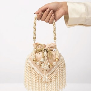 May include: A cream-colored satin drawstring handbag featuring an intricate exterior adorned with rows of hanging white pearl fringe, gold-toned metallic accents, and a braided handle embellished with pearls and gold thread detailing.