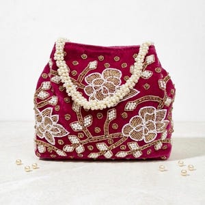 May include: A burgundy velvet handbag with a beaded handle and floral embroidery. The bag features gold and white beaded floral designs and scattered pearls. A stylish accessory for special occasions.