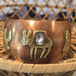 May include: A wide copper cuff bracelet featuring a central brass donkey inlay with an iridescent abalone shell saddle. The bracelet is decorated with additional brass cutouts of a cactus on the left and a stylized figure on the right side.