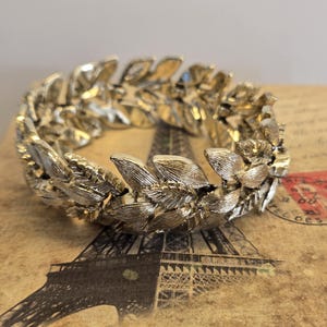 May include: A gold-toned metal bracelet in a laurel wreath design. The bracelet is circular, with detailed leaf and flower elements. It rests on a vintage postcard with an Eiffel Tower illustration and French text.