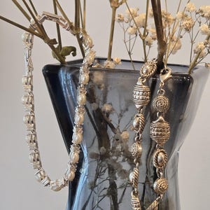 May include: Two silver-toned bracelets. The left bracelet features a series of round white pearls alternating with small clear stones in a metal setting. The right bracelet consists of textured, patterned silver-toned metal links.