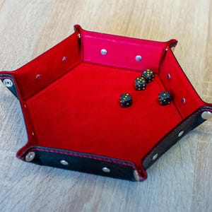 May include: A red and black leather dice tray with a hexagonal shape. The interior is bright red, and the exterior is black with silver rivets. Four black dice with yellow dots are inside the tray.