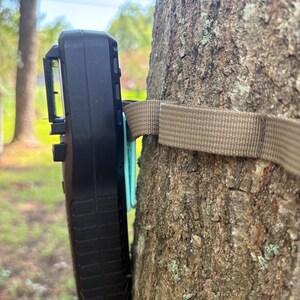 May include: A black trail camera is mounted on a tree trunk with a brown strap. The camera has a rectangular shape with a textured surface and a clip on the side. The tree bark is brown with patches of green.