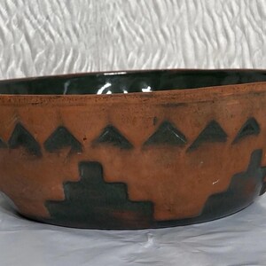 May include: A round terracotta bowl featuring a dark green interior and a matching dark green geometric pattern on the exterior. The design consists of a row of triangles near the rim and a stepped, pyramid-like motif around the base.