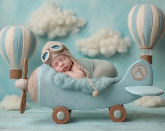 May include: A sleeping newborn, wearing a pilot's hat and goggles, is nestled in a light blue wooden toy aeroplane. The scene is set with fluffy clouds and hot air balloons in the background, creating a whimsical atmosphere.