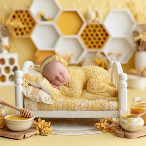 Puede incluir: Un bebé dormido con un conjunto y una diadema de punto amarillo descansa sobre una cama blanca en miniatura con una manta estampada amarilla. Los accesorios con temática de miel incluyen un cuenco de miel, un cucharón de miel y un tarro de miel, todo sobre un fondo amarillo.