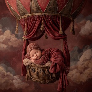 Puede incluir: Un bebé recién nacido durmiendo en una cesta de mimbre suspendida de un globo aerostático rojo y marrón. El bebé está envuelto en una manta y un gorro rojos a juego. El fondo presenta un cielo nublado y rojizo.