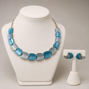May include: A necklace and earring set featuring alternating curved, smooth segments in shades of bright blue and soft gray. The necklace is constructed with a silver-toned metal chain, and the matching earrings consist of two curved pieces.