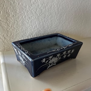 May include: A rectangular ceramic planter with a deep navy blue glaze and white hand-painted floral motifs along the exterior sides. The interior of the basin features a light blue mottled finish, and the base is supported by four small feet.