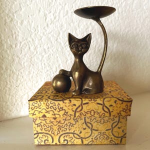 Puede incluir: Una figura de gato de metal en tono bronce que muestra un felino estilizado sentado con una pata apoyada sobre una pequeña esfera. Una cola larga y curva se extiende hacia arriba para sostener un plato circular poco profundo sobre una caja amarilla.