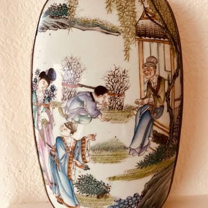 May include: An oval-shaped, antique porcelain container with a hand-painted scene of figures in traditional Asian attire. The artwork features a detailed landscape with trees, a small building, and figures in various poses, all set against a white background.